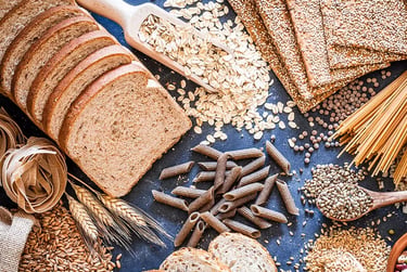 Selection of whole grain foods including sliced bread, pasta, oats, and seeds on a dark surface.