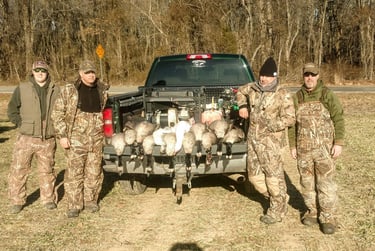 Four hunters in camouflage standing behind a truck tailgate displaying a harvest of Canada geese in a grassy field.