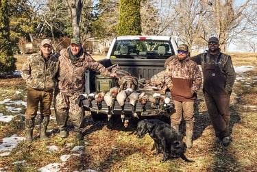 A hunting party of five standing with their black lab behind a truck tailgate full of harvested birds.
