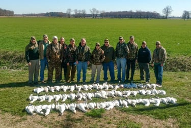 Ten hunters in various outdoor gear posing behind four long rows of harvested snow geese in a green Maryland field.
