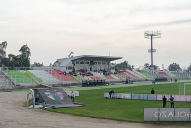 Vista lateral da arquibancada do Estádio Municipal de La Cisterna sob céu azul, com gramado e redes de proteção.