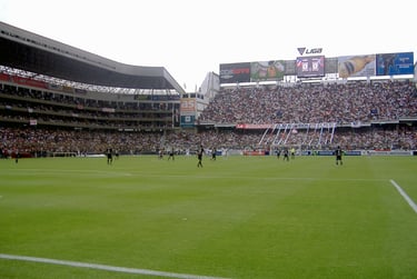 Vista interna do Estádio Rodrigo Paz Delgado em Quito, com arquibancadas brancas e gramado durante uma partida.
