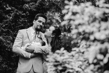 Bride and groom standing among trees with a baby.