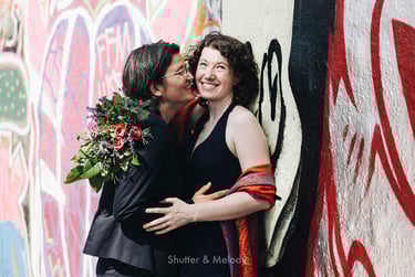 Two brides cuddling next to a wall with graffiti.