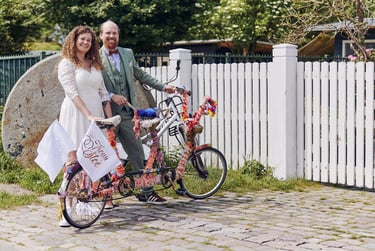 Bride and groom posing next to a tandem bicycle next to a white fence.