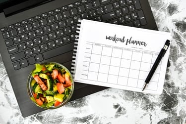 A spiral workout planner and fresh garden salad bowl placed on a laptop keyboard for fitness tracking.
