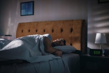 A man sleeping peacefully in a dimly lit bedroom with a digital alarm clock showing 5:59 AM.