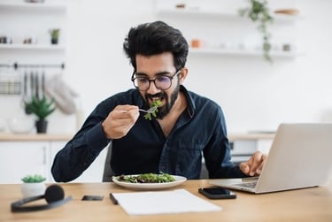 A bearded man wearing glasses eating a healthy salad while working on a laptop at home.