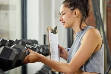 A female personal trainer holding a clipboard while organizing dumbbells on a gym weight rack.