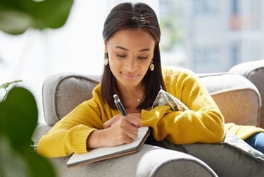 A young woman sitting on a couch writing in a notebook with a pen while relaxing at home.