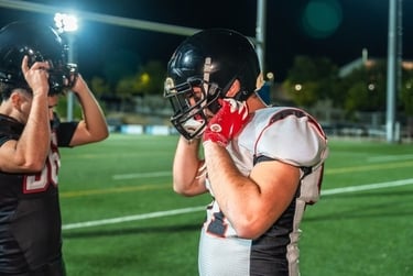 American football players in jerseys and helmets preparing for a night game on a lighted turf field.