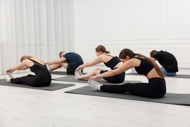 A group of people in a fitness studio performing seated forward fold yoga stretches on mats.