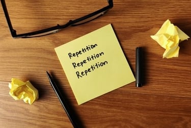 Yellow sticky note with the word repetition handwritten on a wooden desk with a pen and glasses.