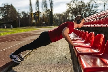 A man in a red shirt performs incline pushups on stadium seats during an outdoor workout.