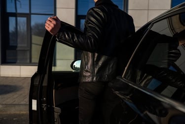 A man in a black leather jacket opens the door of a black luxury car parked outside.