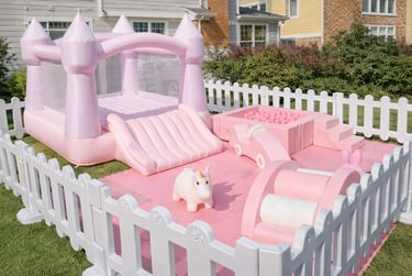 Pink inflatable bounce house with slide and ball pit inside a white picket fence on grass.