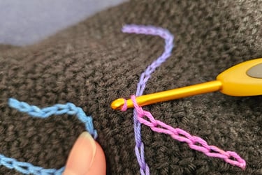 Close-up of a yellow crochet hook weaving colorful pink and purple yarn through black fabric.