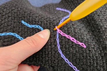 A yellow crochet hook making surface slip stitches with colorful yarn on black fabric.