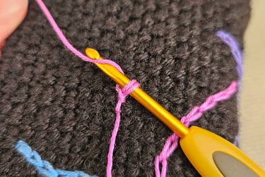 A yellow crochet hook making pink surface slip stitches on a dark brown textured fabric.