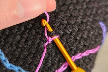 Close-up of a hand using a gold crochet hook to surface crochet pink yarn onto black fabric.