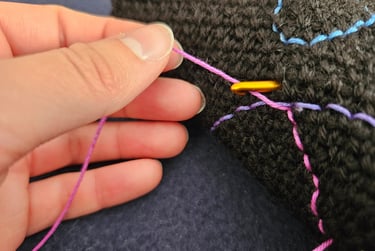 Hand holding pink thread and golden crochet hook to embroider details on a black crochet fabric.