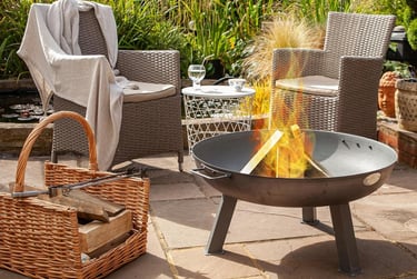 A portable steel fire pit burning logs on a stone patio with wicker garden chairs and a wood basket.