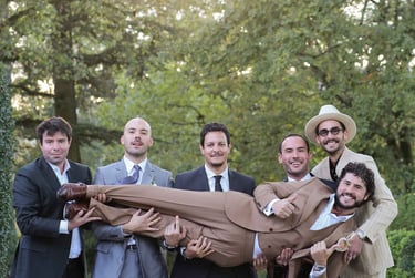 Groomsmen in formal suits playfully lift the groom during an outdoor wedding celebration.