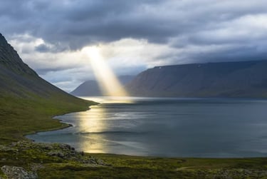 Sunlight breaks through clouds over a peaceful valley and water, showing divine presence in nature.