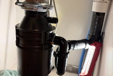 Under-sink view of a new black garbage disposal installation with PVC plumbing and a red work light.