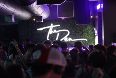 Crowd dancing at The Den nightclub under a white neon sign on a green foliage wall with purple lighting.