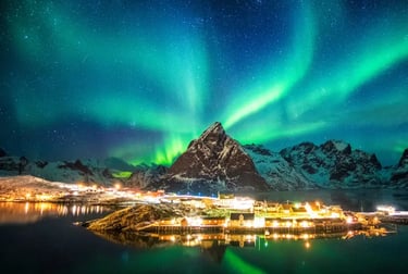 Vibrant green Northern Lights aurora borealis over a snowy mountain and lit village in Reine, Lofoten Islands.