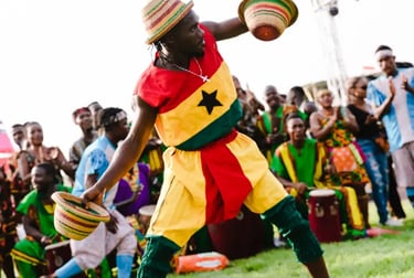 A Ghanaian performer in a traditional flag costume dances with woven hats during a cultural festival.