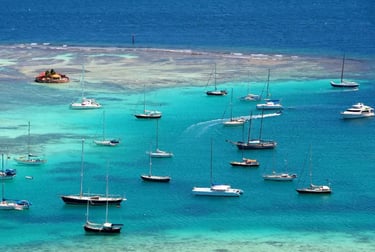 Aerial view of sailboats and catamarans anchored in the turquoise Caribbean waters of a tropical island bay.