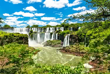 Panoramic view of Iguazu Falls with cascading waterfalls and lush green tropical rainforest under a blue sky.