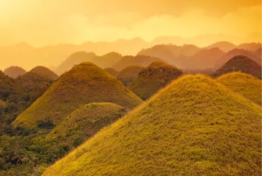 Golden Chocolate Hills of Bohol, Philippines, under a warm sunset sky with misty mountains.
