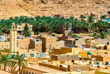 Scenic view of a traditional desert town in Algeria featuring a tall minaret, palm trees, and tan buildings.