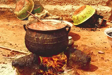 A large black cast iron pot simmering with stew over an open wood fire in a rural outdoor kitchen.