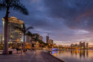 Sunset over Luanda Bay skyline with illuminated buildings and palm trees along the waterfront promenade.