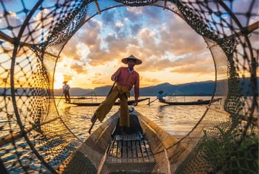 Intha fisherman rowing a wooden boat on Inle Lake at sunset using traditional leg-rowing technique.