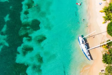 Aerial view of a white sailboat anchored on a tropical beach with turquoise ocean water and palm trees.