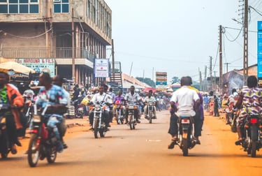 Busy street in Benin with people riding motorcycles and scooters past local market shops.