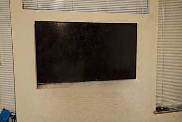 Large flat screen TV mounted on a white wall between windows with horizontal blinds.