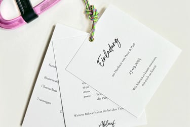 eine schlichte Hochzeitseinladung, bestehend aus mehreren Seiten. Sie wird durch eine Öse und ein Kl