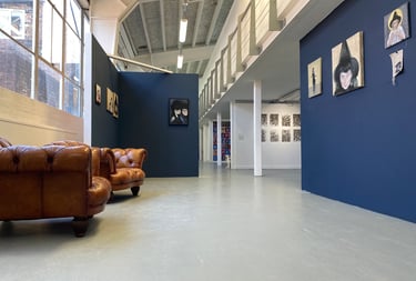 Oil Paintings hung on dark blue walls in a large gallery. There is a window and seating to the left.