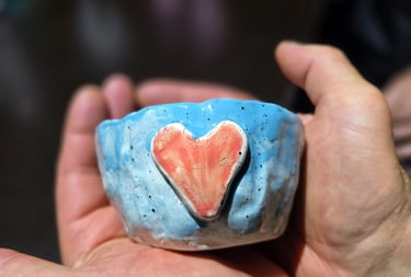 Handmade light blue ceramic bowl with a red clay heart being held in a pair of open hands.
