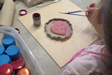 A young child painting a pink ceramic heart dish with a brush during a pottery craft workshop.