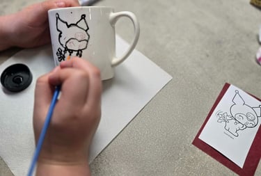 A person hand-painting a cartoon character onto a white ceramic coffee mug with a blue paintbrush.