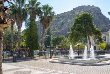 A sunny public square in Nafplio featuring a water fountain, palm trees, and a view of Palamidi Fortress.