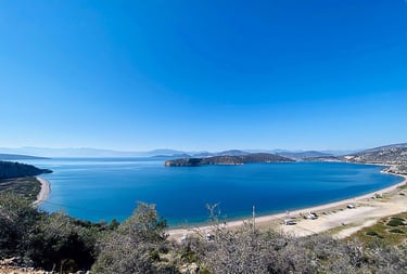 Aerial view of a scenic blue bay and sandy beach in Greece with mountains under a clear sky.