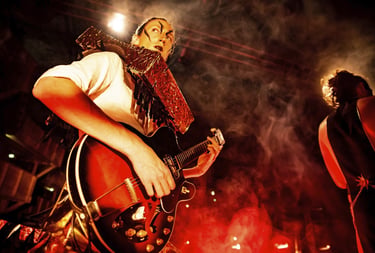 A theatrical guitarist with face paint plays a hollow body electric guitar on a smoky stage.